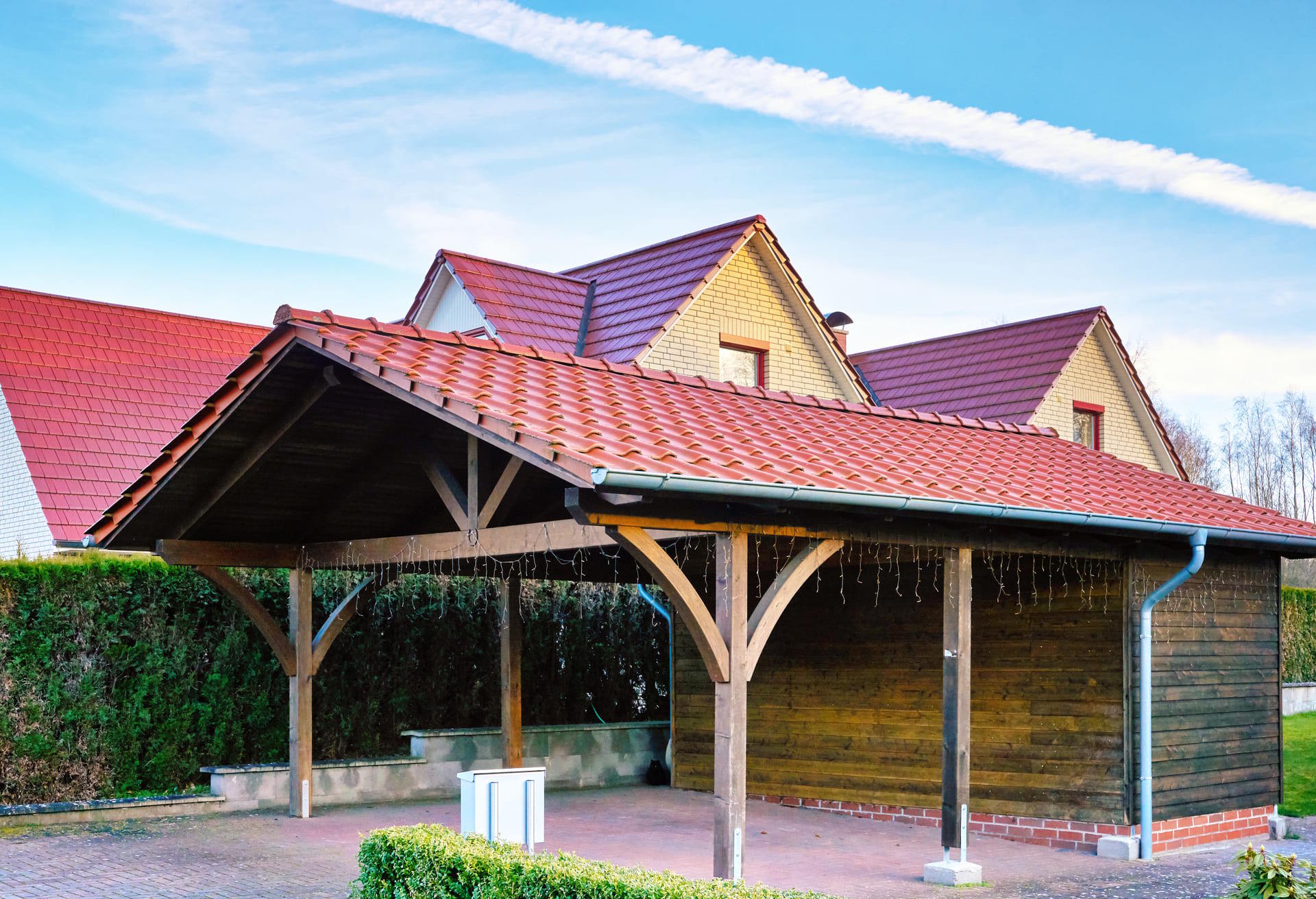 Carport aus Holz mit Satteldach und roter Eindeckung neben einem Einfamilienhaus