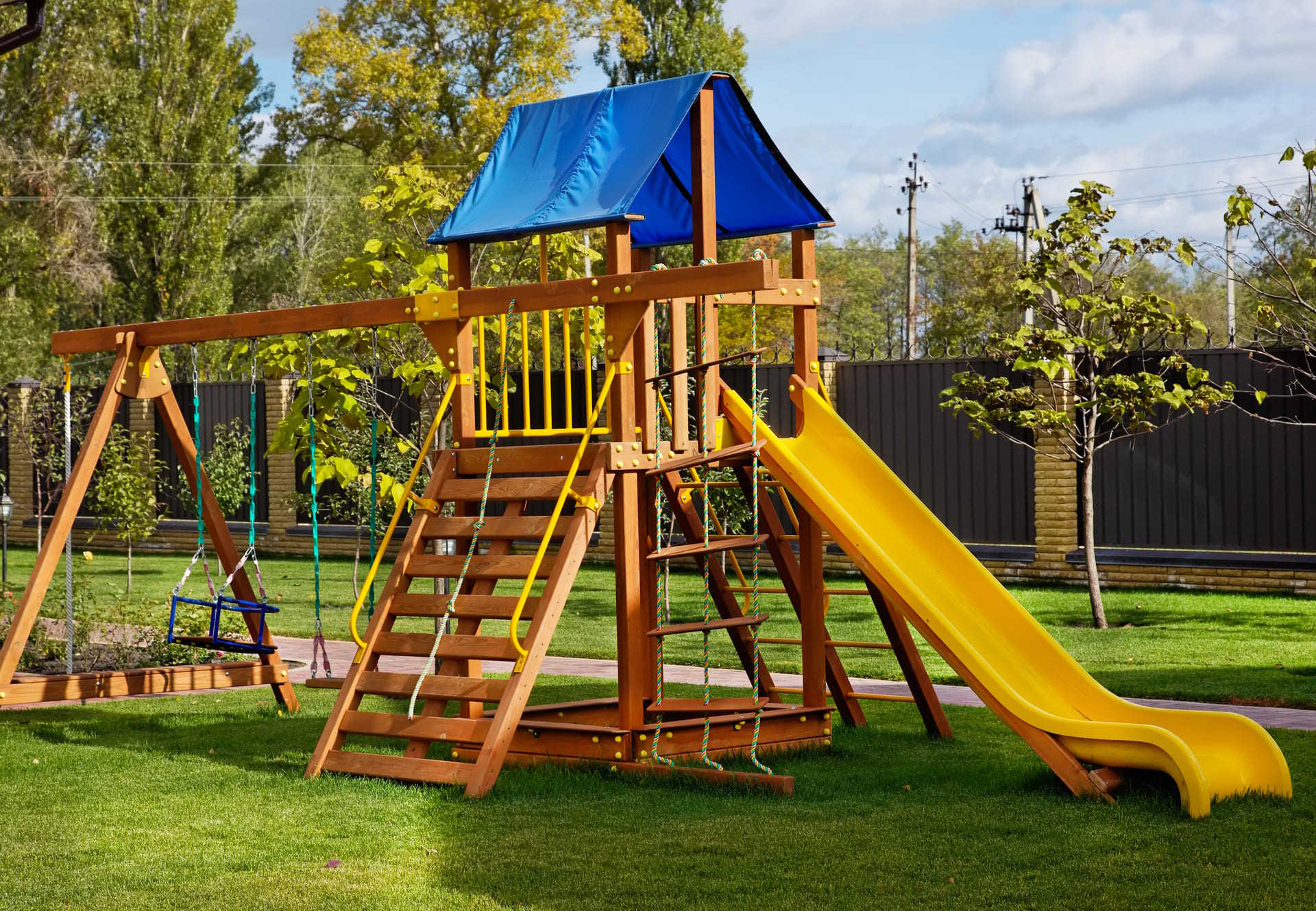 Spielturm aus Holz mit Dach, gelber Rutsche und Schaukel auf einer Rasenfläche